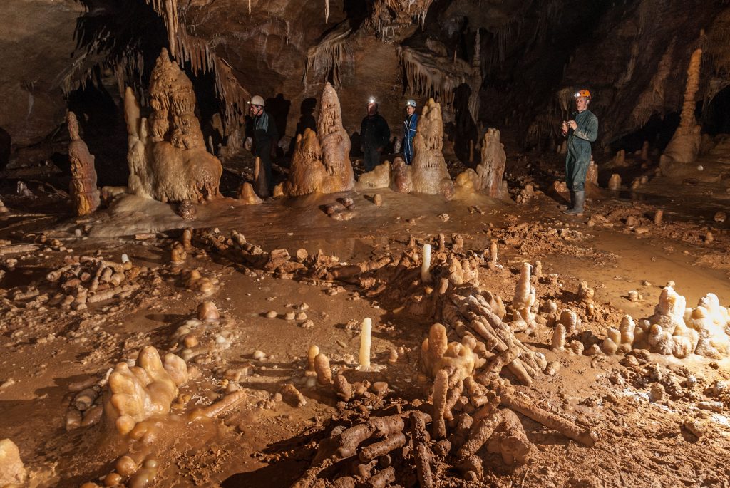 A photograph features four people with headlamps inside a large cave with brown rocky ground and several stalagmites and stalactites.