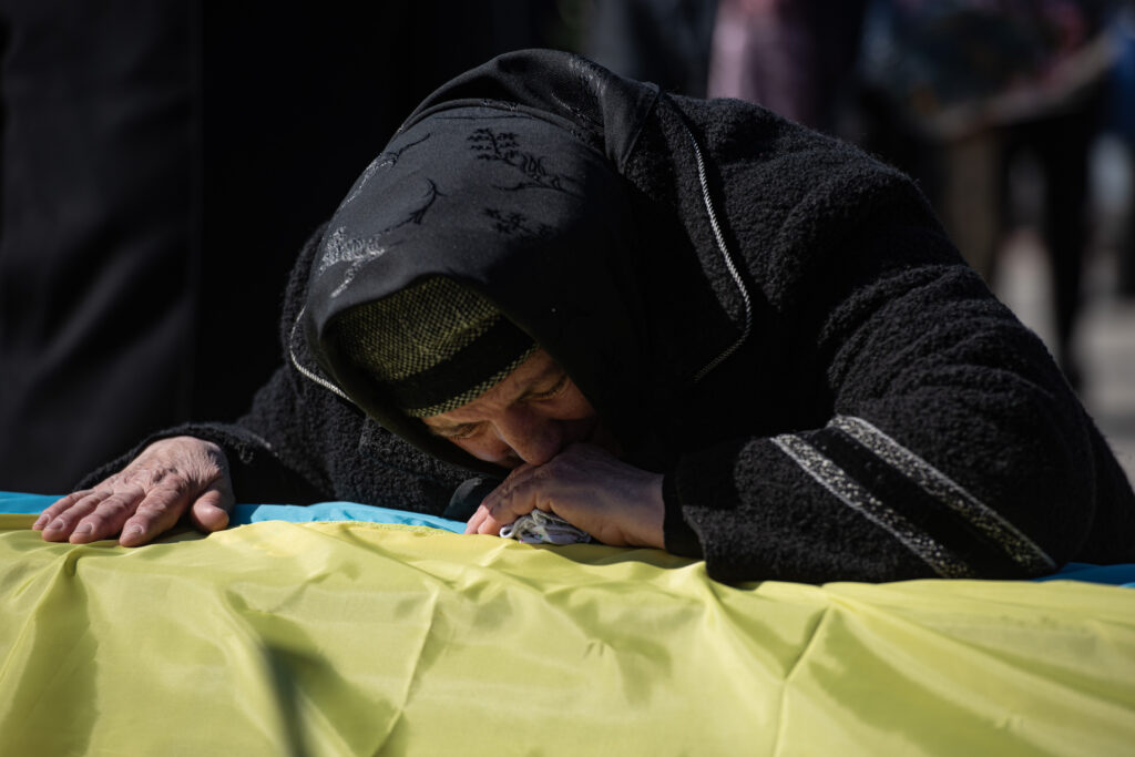 A close-up photograph features a person in a black headwrap and dress crying into their hand as they lean on a coffin covered with a blue-and-yellow flag.