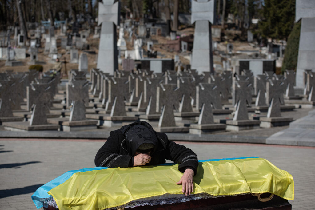 A photograph features a person leaning on a coffin covered with a blue-and-yellow flag. Numerous gray tombstones are in the background.