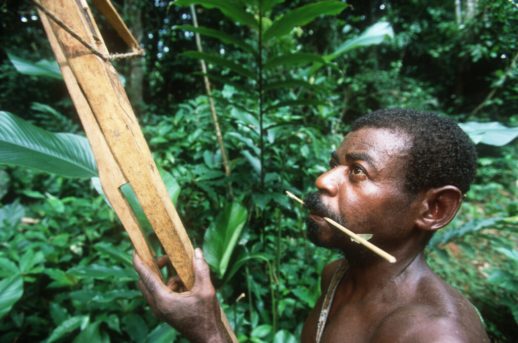 A close-up photograph features a person with short black hair and a mustache holding an arrow fastened to a stick in their mouth while grasping a wooden hunting tool.