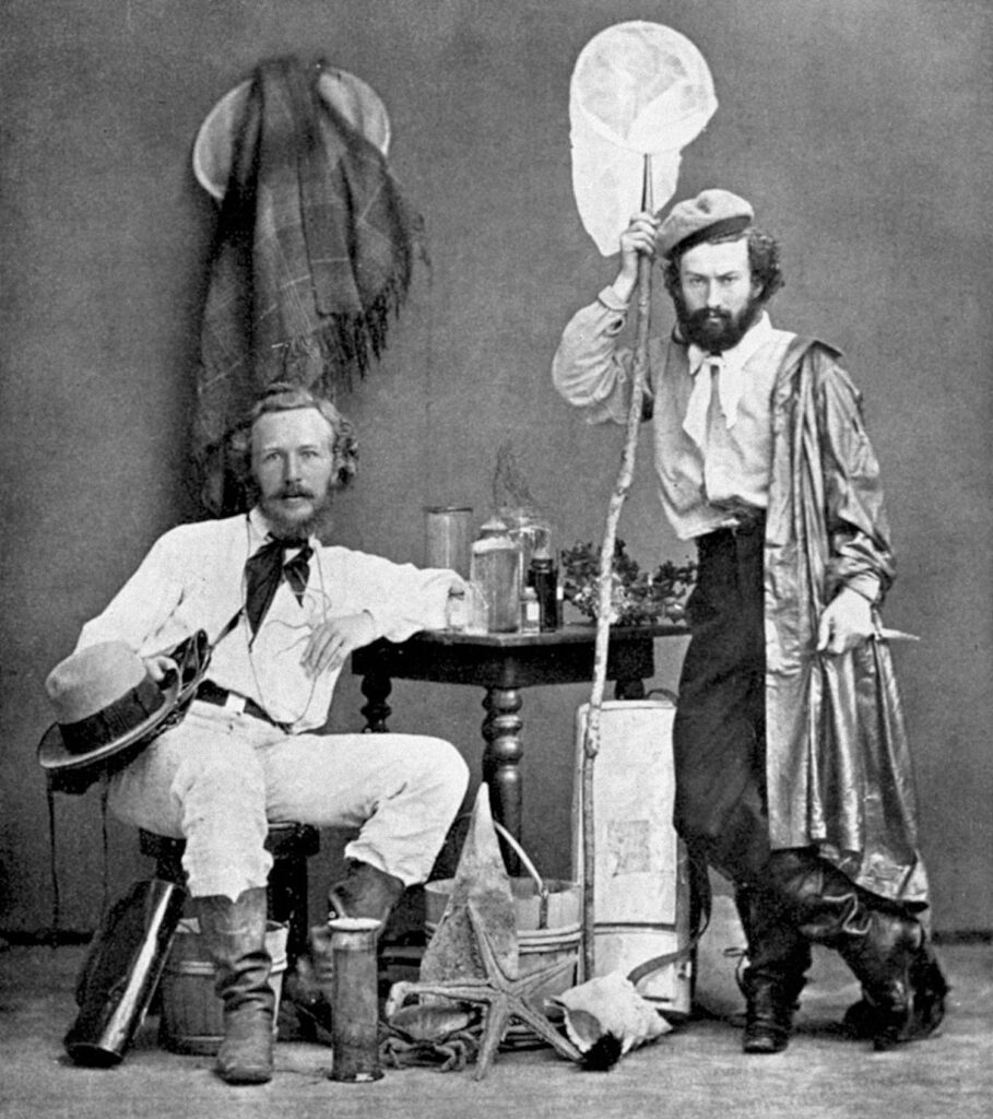 A black-and-white archival photograph features two people posing near a table with various objects on it. The seated person on the left balances a hat on his knee. The other person stands and holds an insect net.