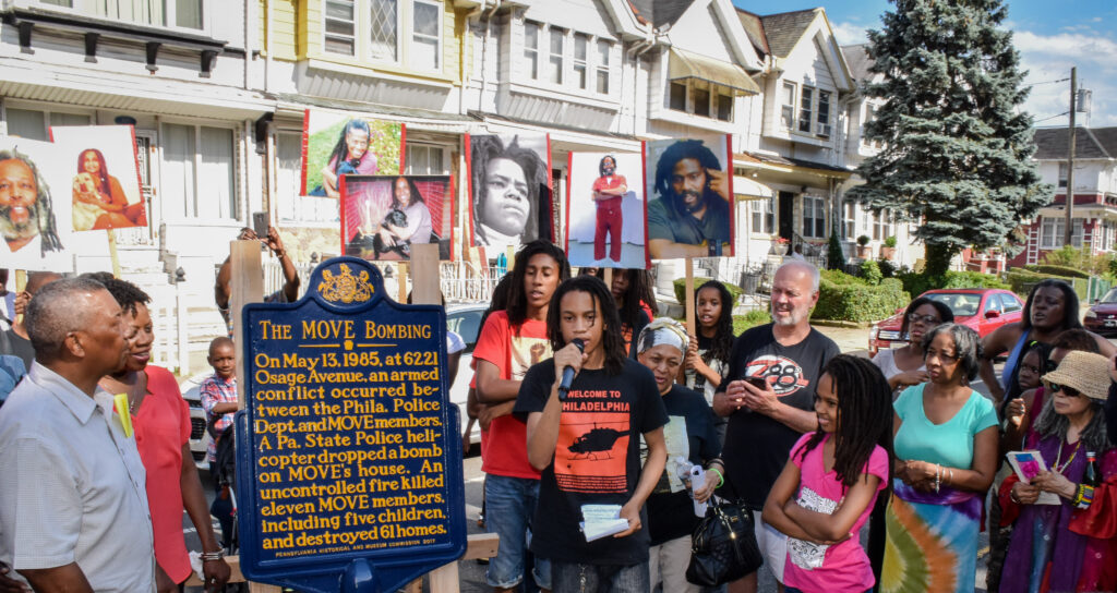 A photograph features a crowd around a blue sign. One person speaks into a microphone and several hold of photos of people’s faces. The sign reads, “The MOVE Bombing: On May 13, 1985, at 6221 Osage Avenue, an armed conflict occurred between the Philadelphia Police Department and MOVE members. A Pennsylvania State Police helicopter dropped a bomb on MOVE’s house. An uncontrolled fire killed 11 MOVE members, including five children, and destroyed 61 homes.