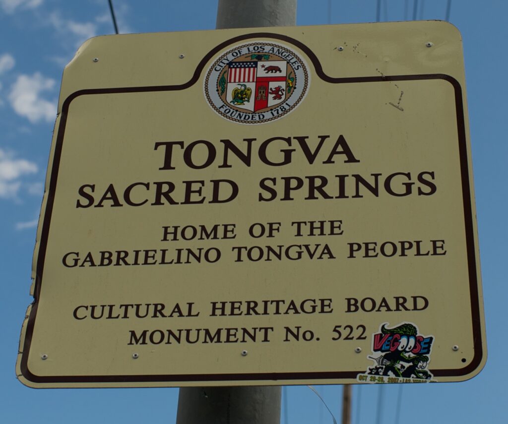 A photograph features a yellow sign on a telephone pole with a crest on top surrounded by a circle of text that reads, “City of Los Angeles, Founded 1781.” Under that, text reads, “Tongva Sacred Springs, home of the Gabrielino Tongva People, Cultural Heritage Board Monument No. 522.”