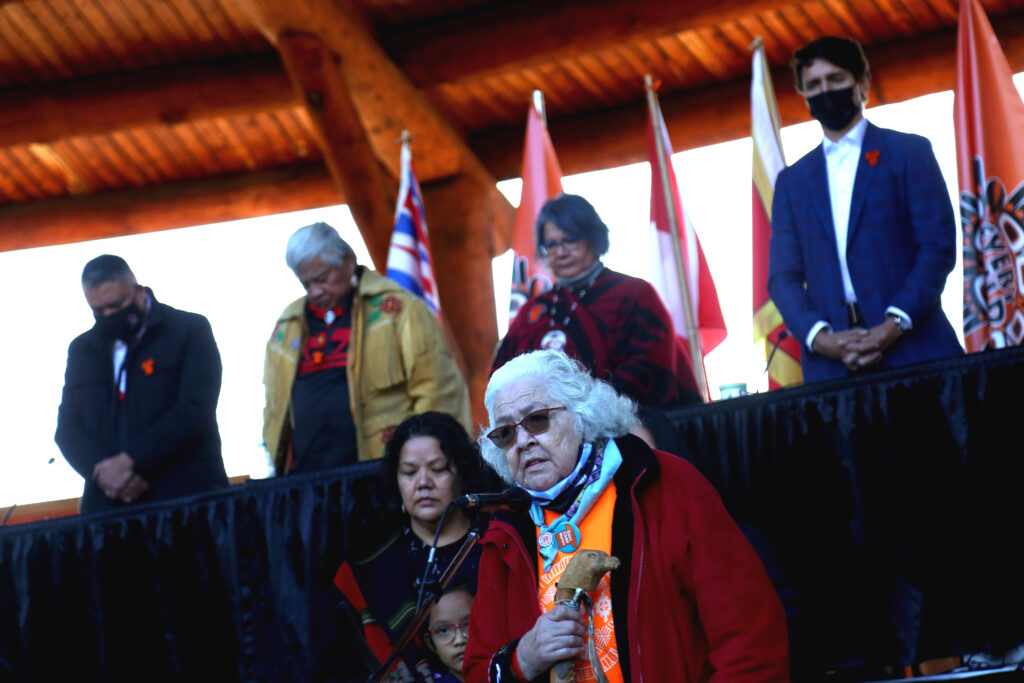 A photograph shows a person with white hair and sunglasses standing in front of another adult and a child. On an elevated platform behind them, people stand in a row and bow their heads.