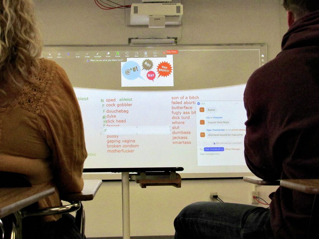 An image shows a projection screen flanked by two people on the left and right. The screen has vertical lists of curse words such as “douchebag,” “smartass,” and “son of a bitch” listed in red font, with a Zoom chat active in the bottom right corner.