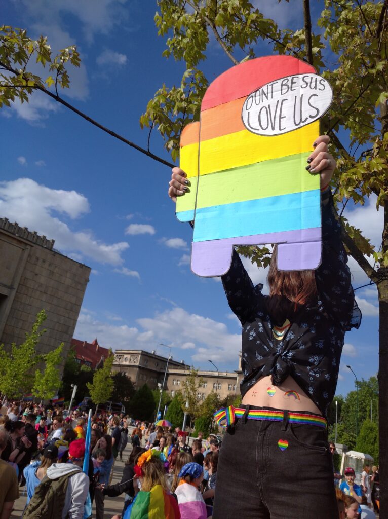 A person in a crowd of rainbow clothes and pride flags holds up a cardboard cutout covered with rainbow stripes and text that reads, “Don’t be sus, Love us.”