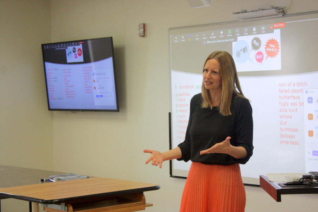 A person with long blonde hair wearing a black shirt and orange skirt gestures in front of a large projection screen and smaller monitor with lists of words. Some words in the lists include “son of a bitch,” “whore,” “slut,” “dumbass,” and “jackass.”