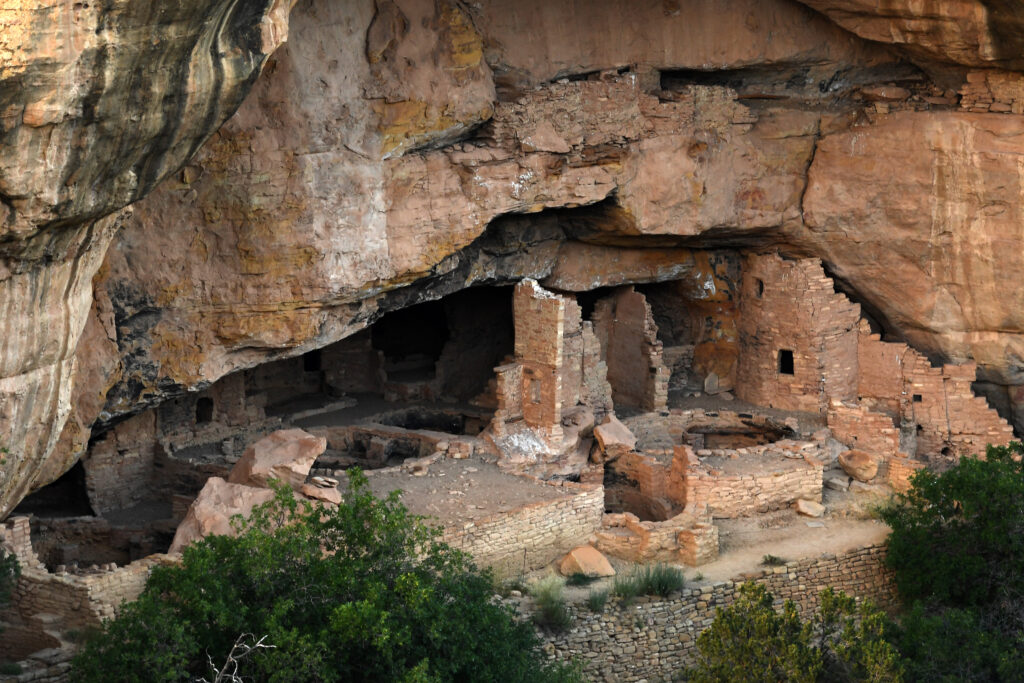 De longe, uma paisagem de estruturas circulares de arenito fica à esquerda de uma parede de arenito com algumas janelas retangulares. Todas as estruturas são construídas em uma grande abertura na lateral de uma parede de penhasco no deserto.