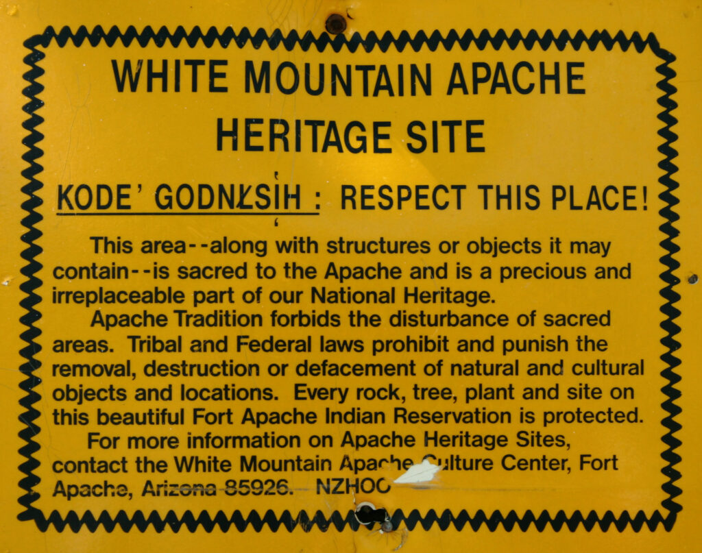 Um sinal amarelo tem uma borda preta ondulada ao redor de um bloco de texto. O cabeçalho diz, em letras maiúsculas, “White Mountain Apache Heritage Site”. Abaixo estão palavras sublinhadas na língua Ndee traduzidas para “Respeite este lugar” em inglês, seguidas por um ponto de exclamação. Abaixo disso, um parágrafo diz: “Esta área – junto com as estruturas ou objetos que ela pode conter – é sagrada para os Apaches e é uma parte preciosa e insubstituível de nosso Patrimônio Nacional. A Tradição Apache proíbe a perturbação de áreas sagradas. As leis tribais e federais proíbem e punem a remoção, destruição ou desfiguração de objetos e locais naturais e culturais. Cada rocha, árvore, planta e local desta bela Reserva Indígena Fort Apache é protegido. Para obter mais informações sobre os locais do patrimônio Apache, entre em contato com o White Mountain Apache Culture Center, Fort Apache, Arizona 85926. NZHOO.”