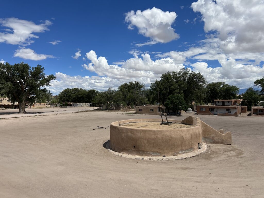 Uma estrutura circular de pedra com uma pequena escada de entrada fica no meio de uma área de terra plana com edifícios retangulares, árvores verdes e um céu azul ao fundo.