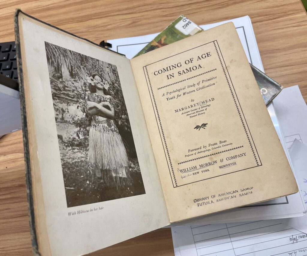 An old, yellowing book is open on a table, with the title page on one side and a young Samoan woman on the other side.