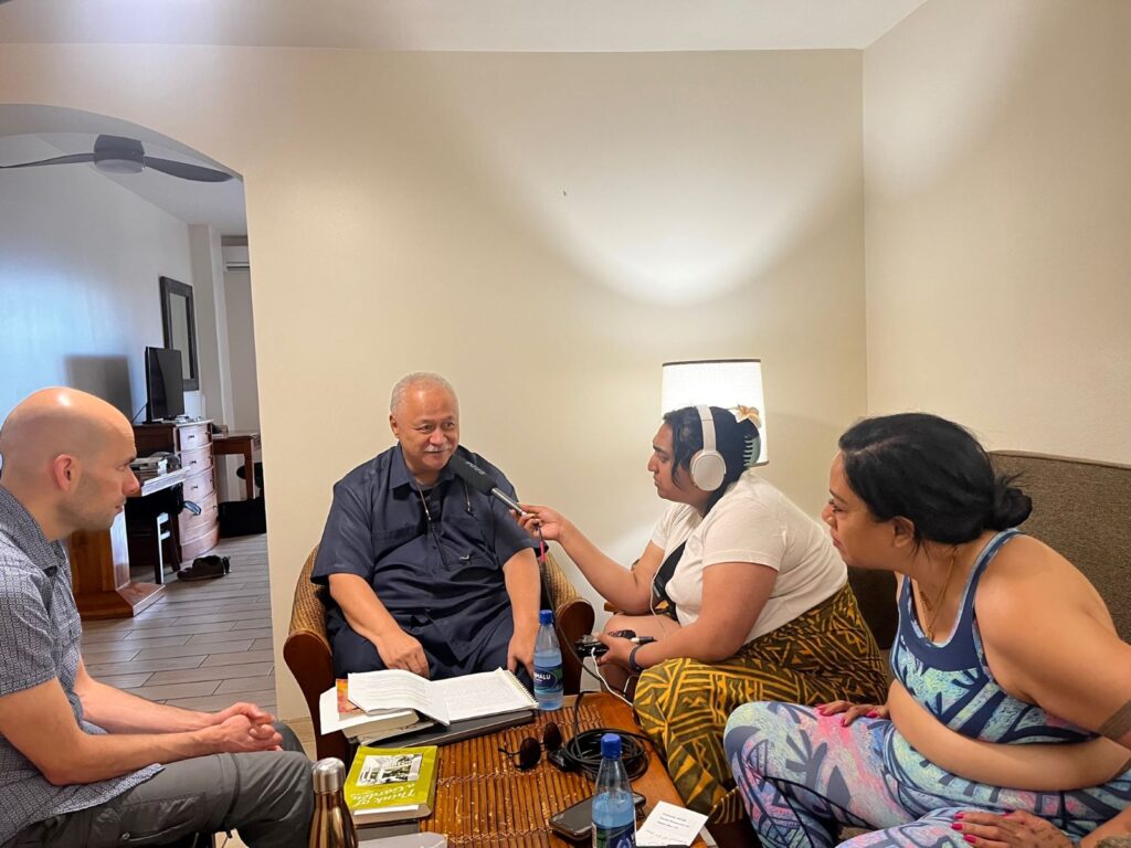 An older man with grey hair and mustache is being interviewed by three people in a plain hotel room.