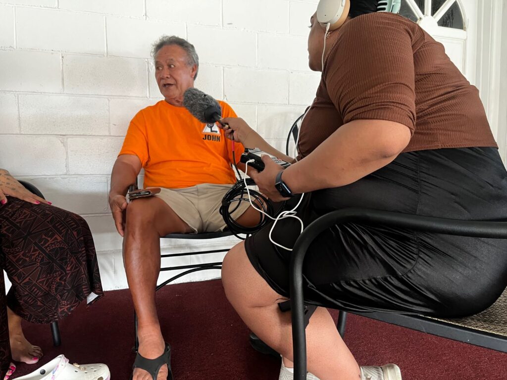 An older man in a bright orange shirt is interviewed by a woman holding a large microphone to him.