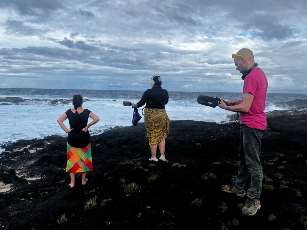 Three people stand on a rocky shore staring out into the ocean, with sound equipment.