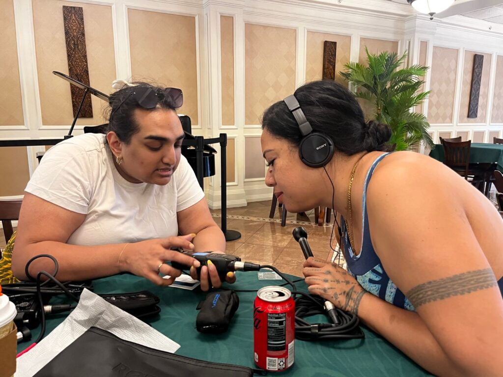 Two women are hunched over a table looking at recording equipment.