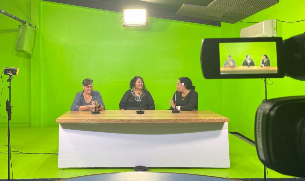 On a TV set three women sit in front of a green painted room and a desk, with a camera recording them off to the right.