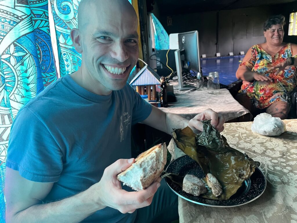 A middle aged man with no hair holds food in one hand and has a plate of food in front of him.