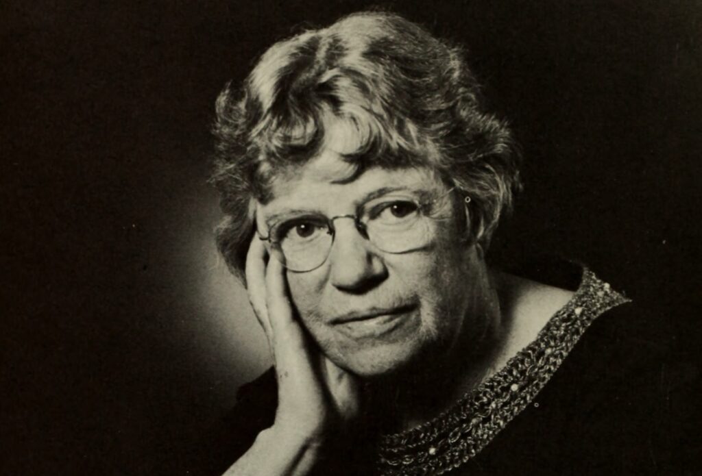 An older white woman poses in a studio. She has short-cropped hair and thick glasses, and has her head leaning gently against one upraised hand.