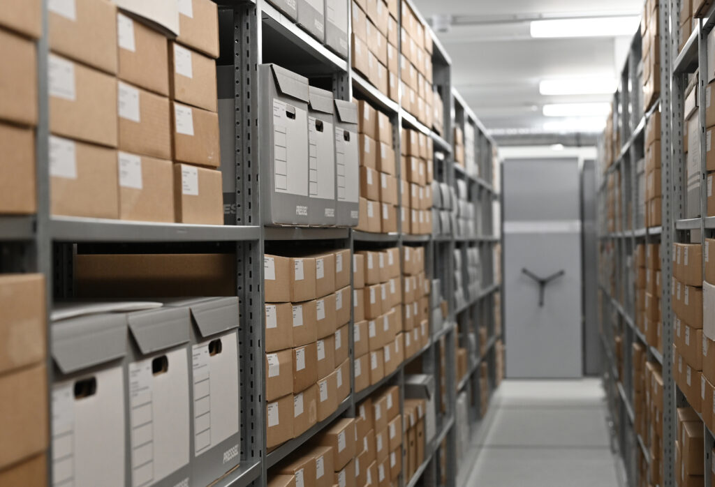 Two metal shelving units on either side of an aisle hold rows of several white and tan cardboard boxes. A small white sticker with text labels each box.