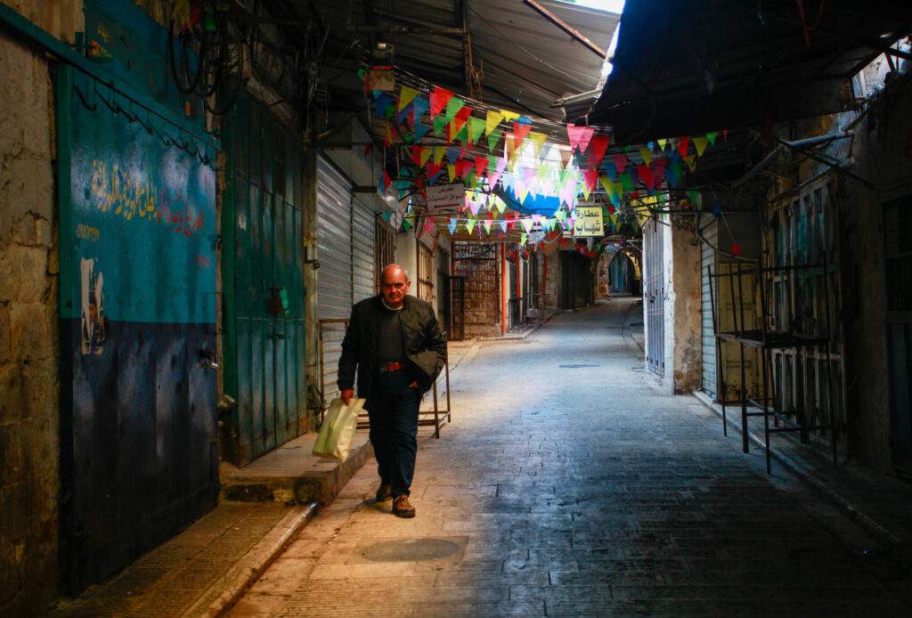 A nearly bald person wearing a coat and carrying a bag walks down a dark covered walkway lined with boarded-up shops. Strings of colorful flags hang overhead.