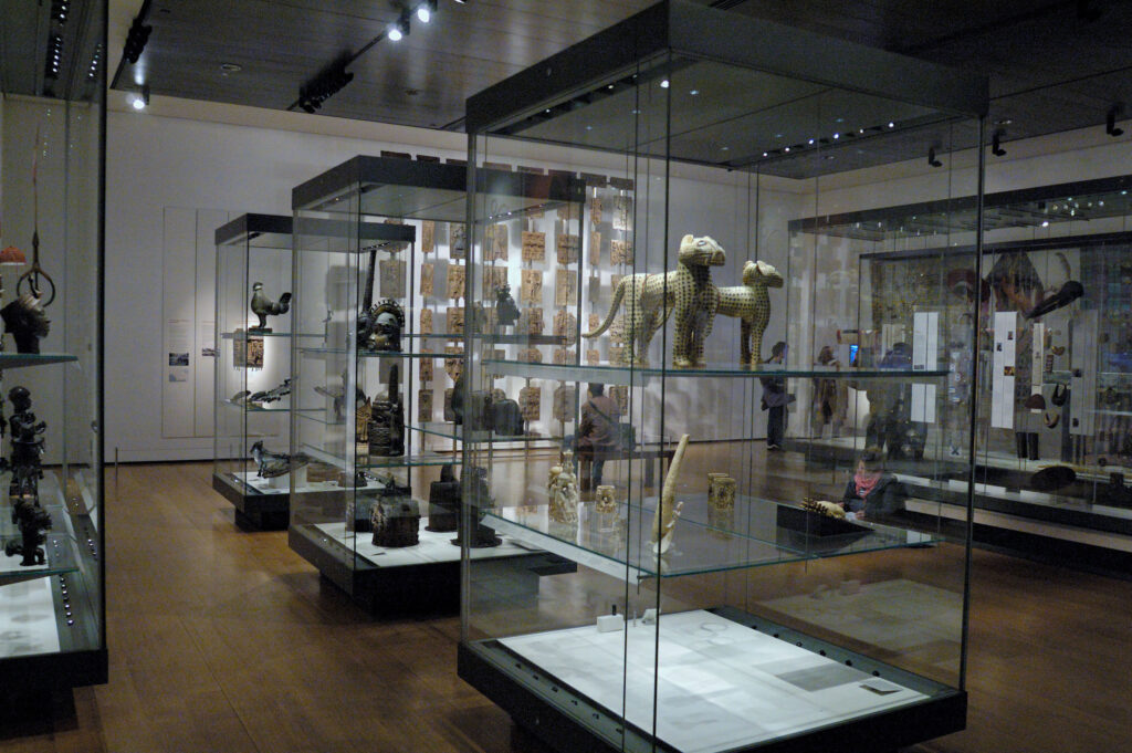 A person crouches on a wooden floor among several glass display cases that hold various objects. Some include carvings of cheetahs, birds, and people wearing ornate headpieces.