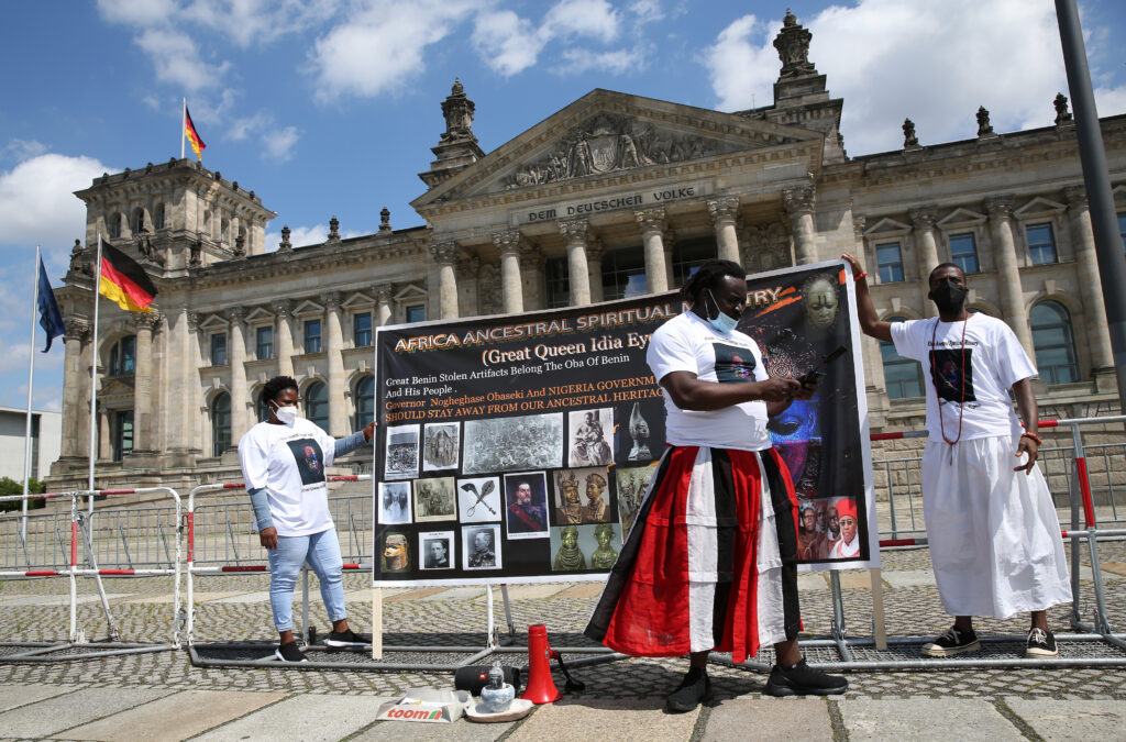 Three dark-skinned people wear white T-shirts with square images on them. Two hold a large poster that reads, Africa Ancestral Spiritual Ministry. Text below says, Great Queen Idia Eyes. Great Benin Stolen Artifacts Belong the Oba of Benin and His People. Governor Nogeghase Obaseki and Nigeria government should stay away from our Ancestral Heritage. Visible behind the frontperson, a colorful mask fills the right side of the poster. Under that are several archival images of objects, groups of African people, and different European men.