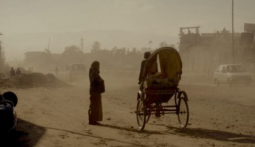 In a dusty, sepia-toned scene, a person wearing a headdress and long dress stands in the middle of a dirt road while a person operating a rickshaw passes them. A building, car, trees, and a distant mountain range fill the background.