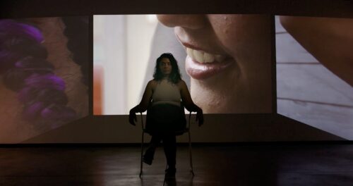 A silhouetted woman sits in a chair in the center of a dark room looking directly at the camera. Close-up images of another woman are projected onto screens on the three walls surrounding her.