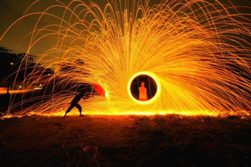 A person stands inside a moving flame of yellow light while sparks fly out in all directions. A second person stands on the left side holding an umbrella against the sparks.