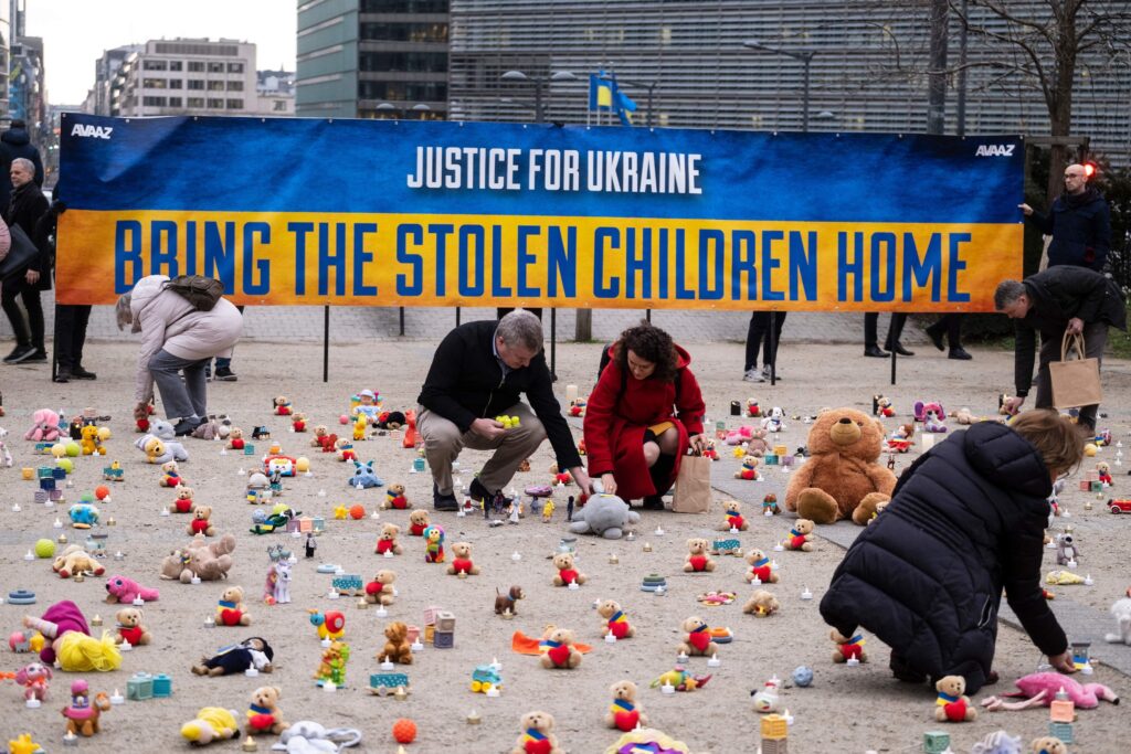 In front of a blue and yellow sign that reads “Justice for Ukraine: Bring the Stolen Children Home,” five people ignite candles next to numerous small teddy bears in a city square.