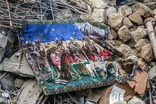A colorful tapestry depicting a traditional scene of Jesus’ birth, with people in robes and headscarves, rests awkwardly on strewn rubble and debris.