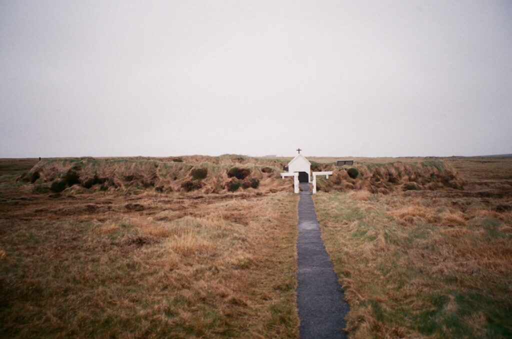 A narrow, paved trail cuts through a grassy field and ends at a small white building in the distance that has a cross on its roof.