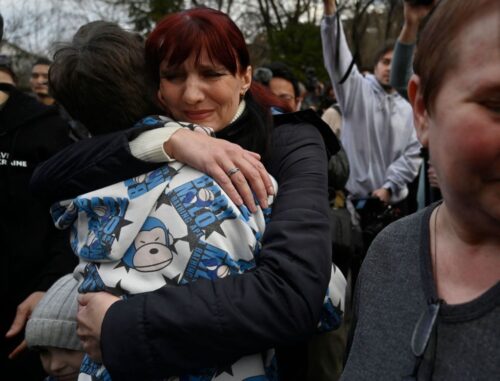Amid a crowd, a woman with dark auburn hair and a black jacket embraces a child who faces away from the camera and wears a jacket with blue cartoon monkeys on it.