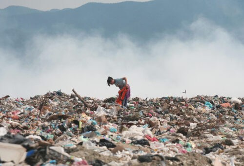 A young person holding a red sack stands atop an enormous heap of trash, backdropped by white smoke from burning garbage and mountains in the distance.