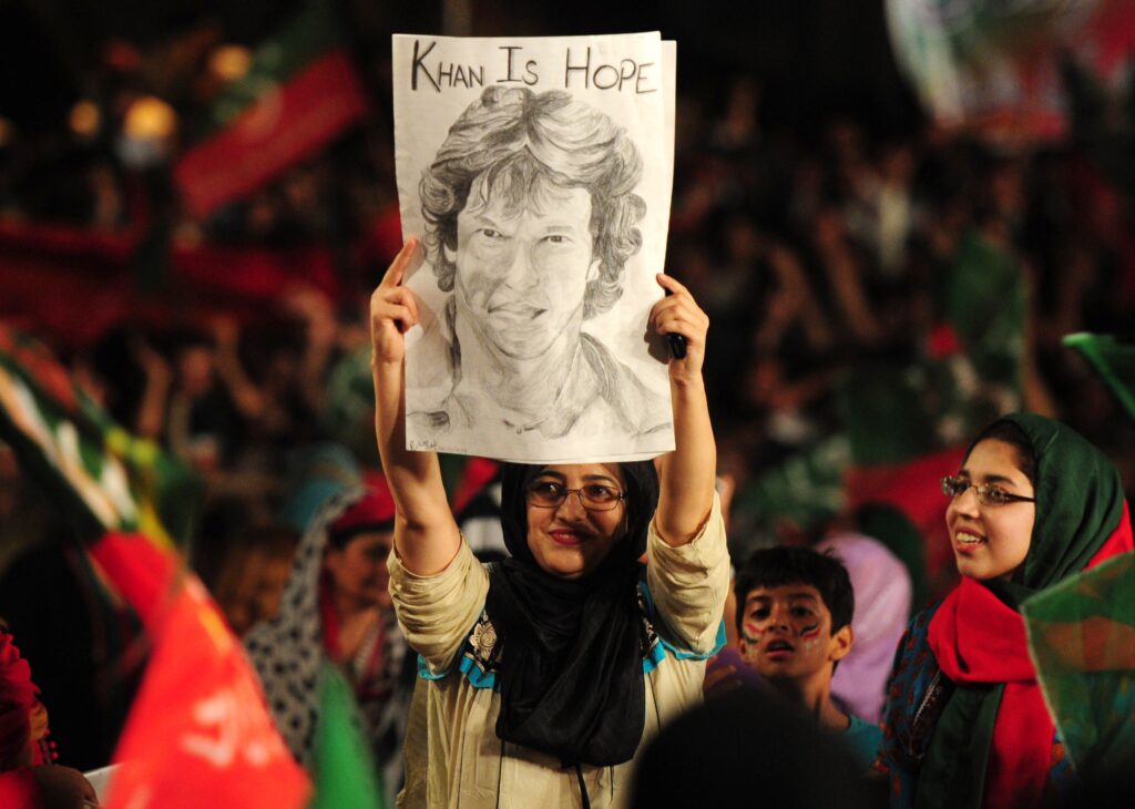 Amid a political rally where red, black, and green colors are prominent, a smiling woman wearing glasses, a khaki shirt, and a black headscarf holds aloft a sketch of a person’s face that says, “Khan Is Hope.”