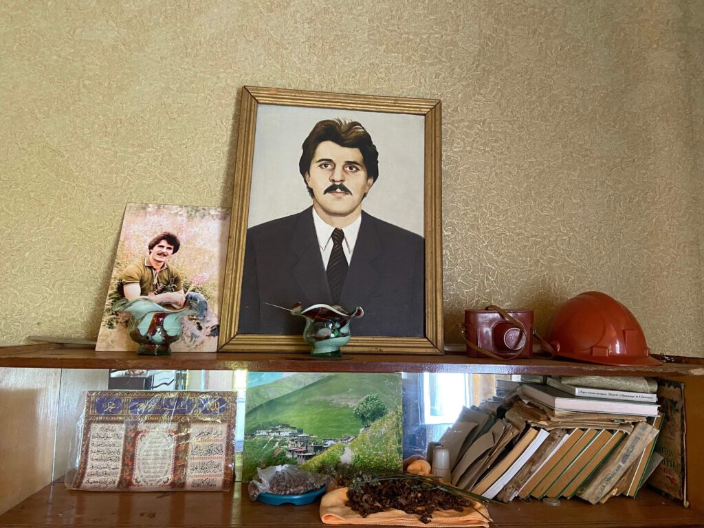 A dark shelf leaning against a wall covered in gold-tinted wallpaper displays the personal items of a missing loved one, including a portrait of the person dressed in a black suit and tie, a photograph, a red plastic hard hat, and books.