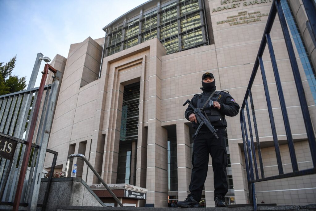 An officer dressed in black, wearing a baseball-style hat and balaclava, with a machine gun slung across his chest, stands before a gray concrete building.