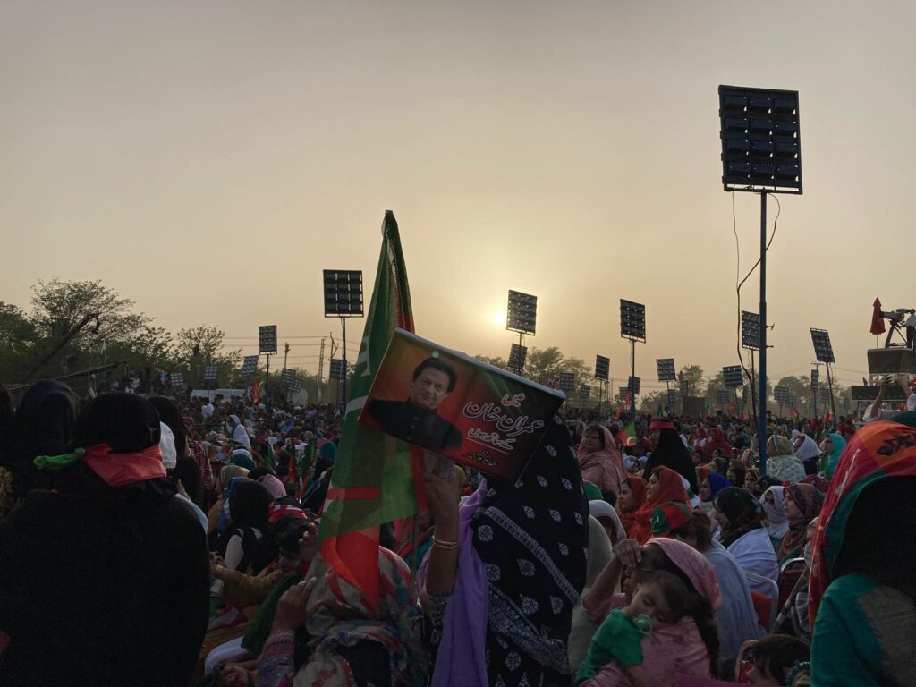 As the sun hangs low in a hazy sky, a crowd bearing red and green flags and political posters stands around an elevated stage surrounded by tall speakers.