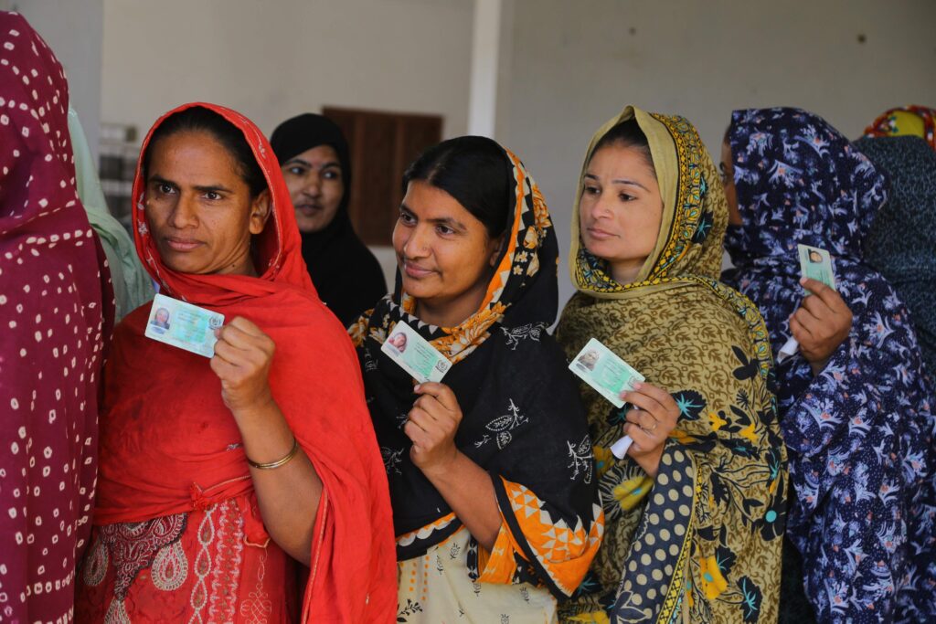 Women wearing brightly colored red, black, orange, yellow, and blue headscarves display their IDs in their left hands as they stand in line.