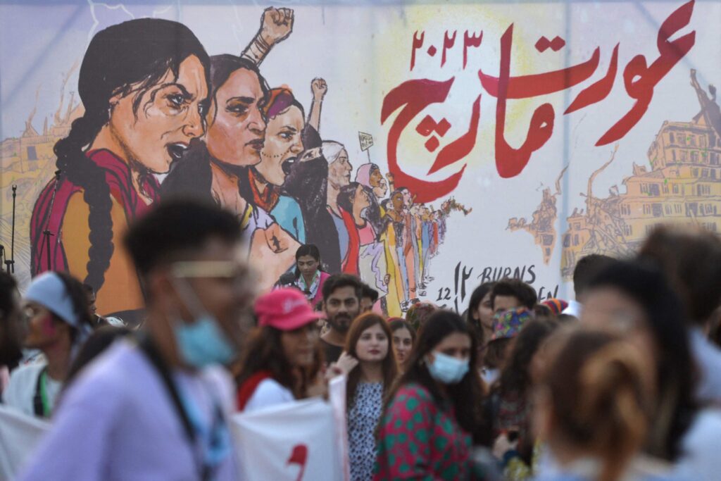 A crowd of demonstrators, some wearing surgical masks, stands in front of a mural depicting politically engaged women with open mouths and raised fists.