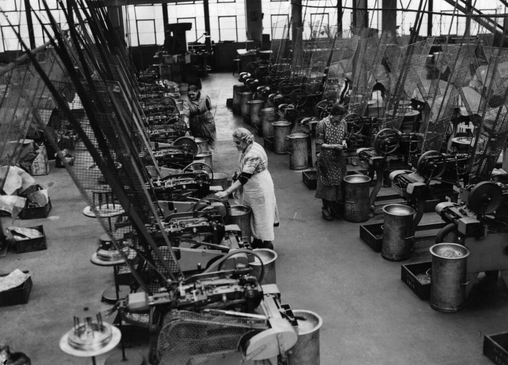 A historic black-and-white photograph shows a small group of workers on the floor of a pin manufacturing plant tending to rows of machines.