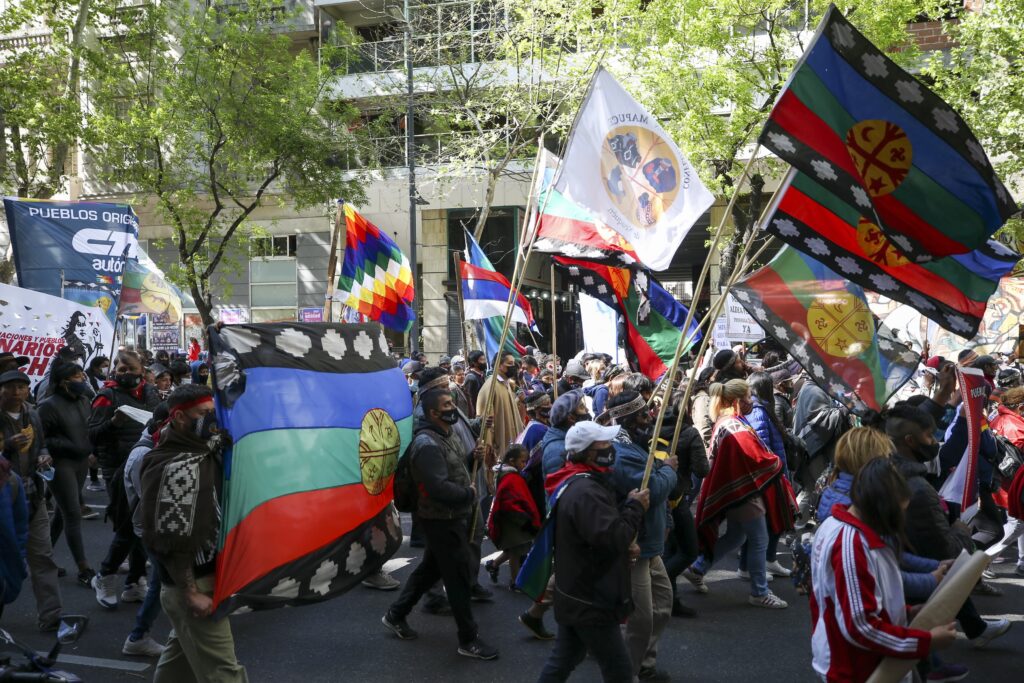 A large group of people walks down a tree-lined street, with some holding colorful flags.