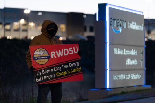 A person wearing a rust-colored hooded coat and a black face mask stands in front of a large facility while holding a red sign that reads: “It’s Been a Long Time Coming but I Know a Change Is Coming!”