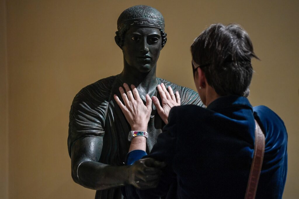 A person in a dark-blue velvet coat stands face to face with a replica of an ancient dark-colored statue of a man and places their hands on its chest.