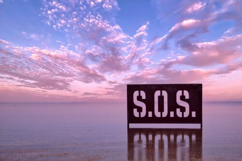 A large metal sign with the letters S.O.S. carved through it stands in a shallow lake in front of a blue sky with pink clouds.