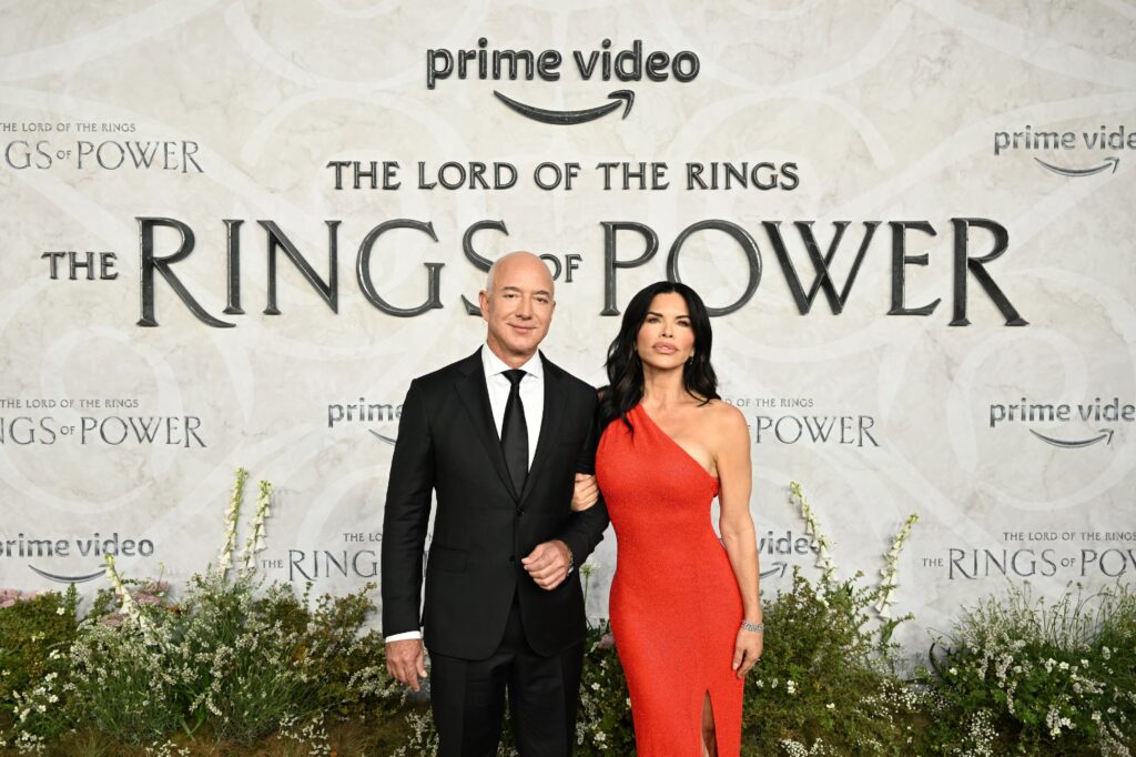 A man and a woman dressed in a black suit and tie and a red dress, respectively, stand arm-in-arm in front of low shrubbery and a large gray backdrop that reads: “The Lord of the Rings: The Rings of Power.”