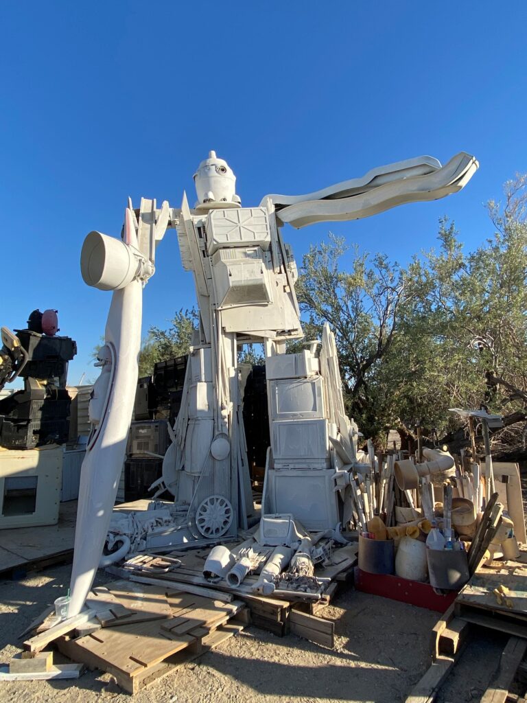 A large robot statue made from discarded equipment and other white-painted objects stands next to old tools, wooden pallets and unidentifiable trash.