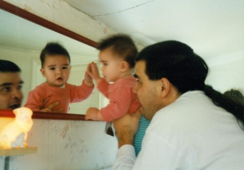 A man with long, dark hair tied in a braid holds up a small child to a mirror, which reflects the child’s pensive face.