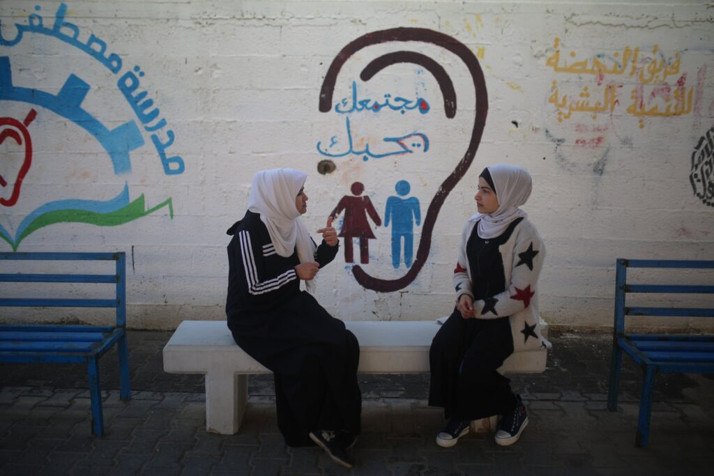 Two people sit face-to-face on a bench and sign with each other. Behind them, a mural shows the outline of a human ear with Arabic writing inside it.