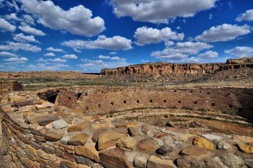 An ancient circular stone structure lies exposed under the ground surface. In the distance, light-red and tan mesas and flat desert expanses lie under a piercing blue sky with scattered fluffy white clouds.